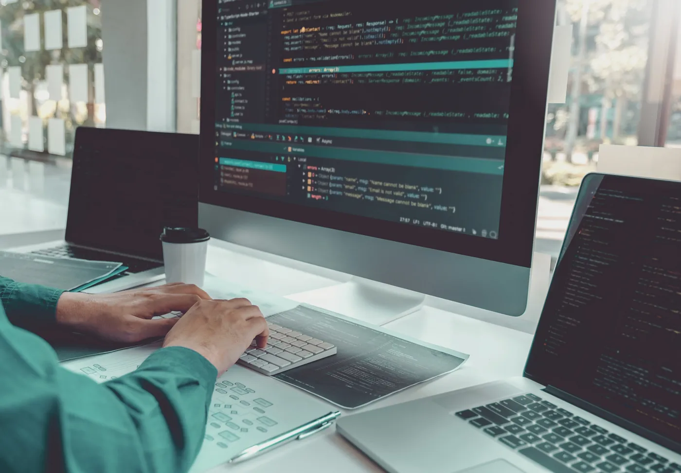 A developer typing at a desk, with code on a large monitor and two laptops beside it.