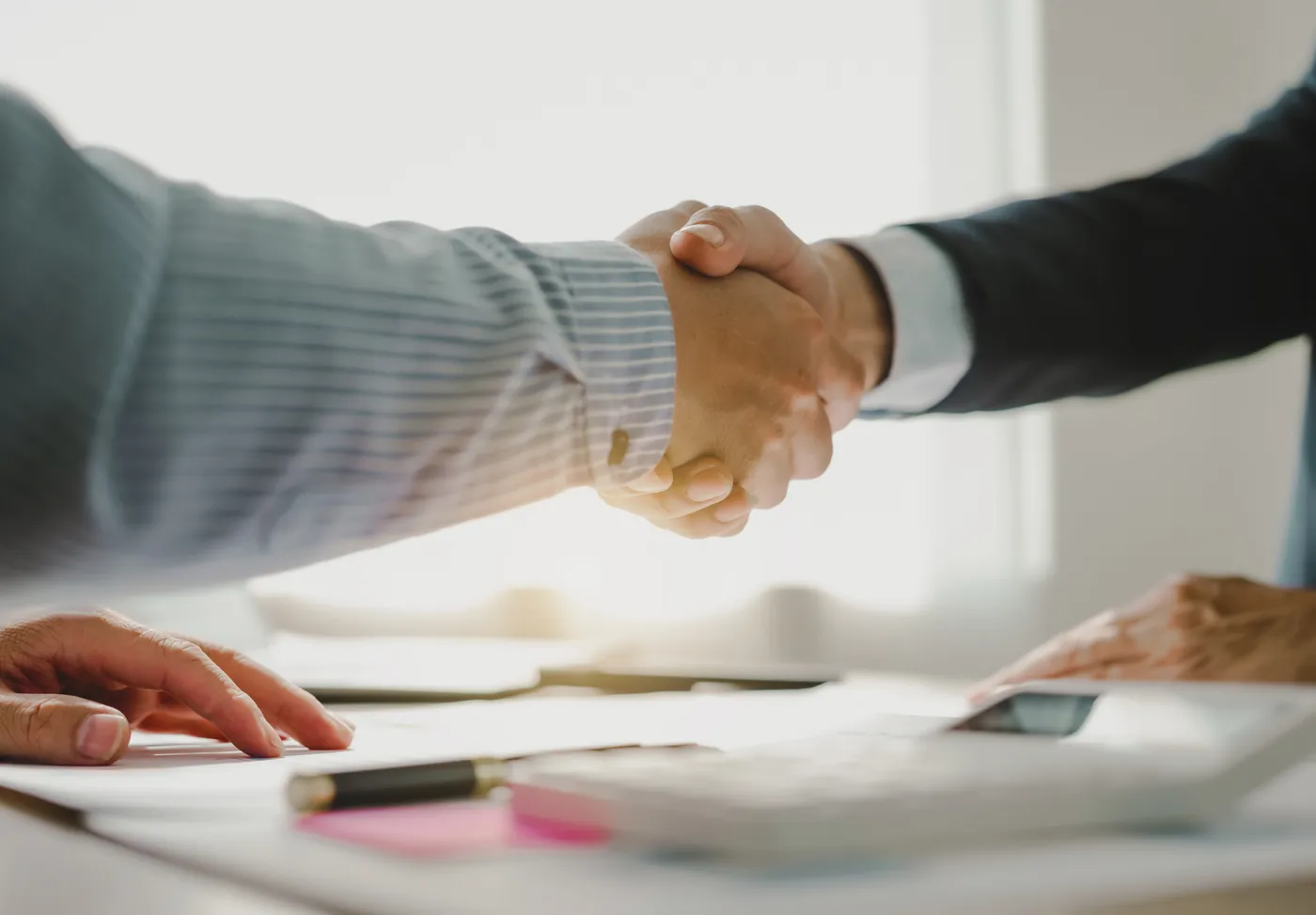 Two people shaking hands across a desk with documents and a calculator.
