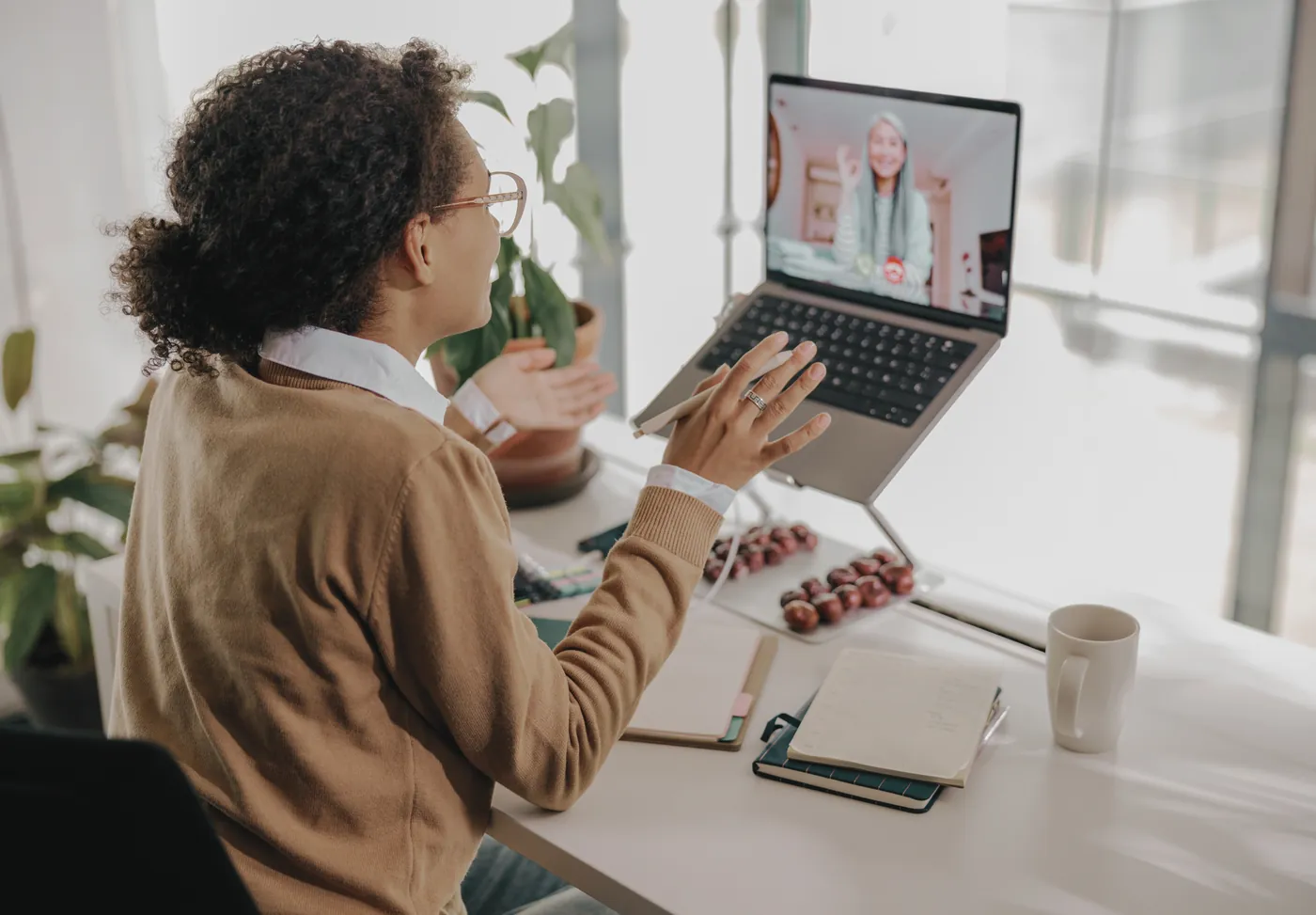 A woman on a video call with a colleague, gesturing towards her laptop.