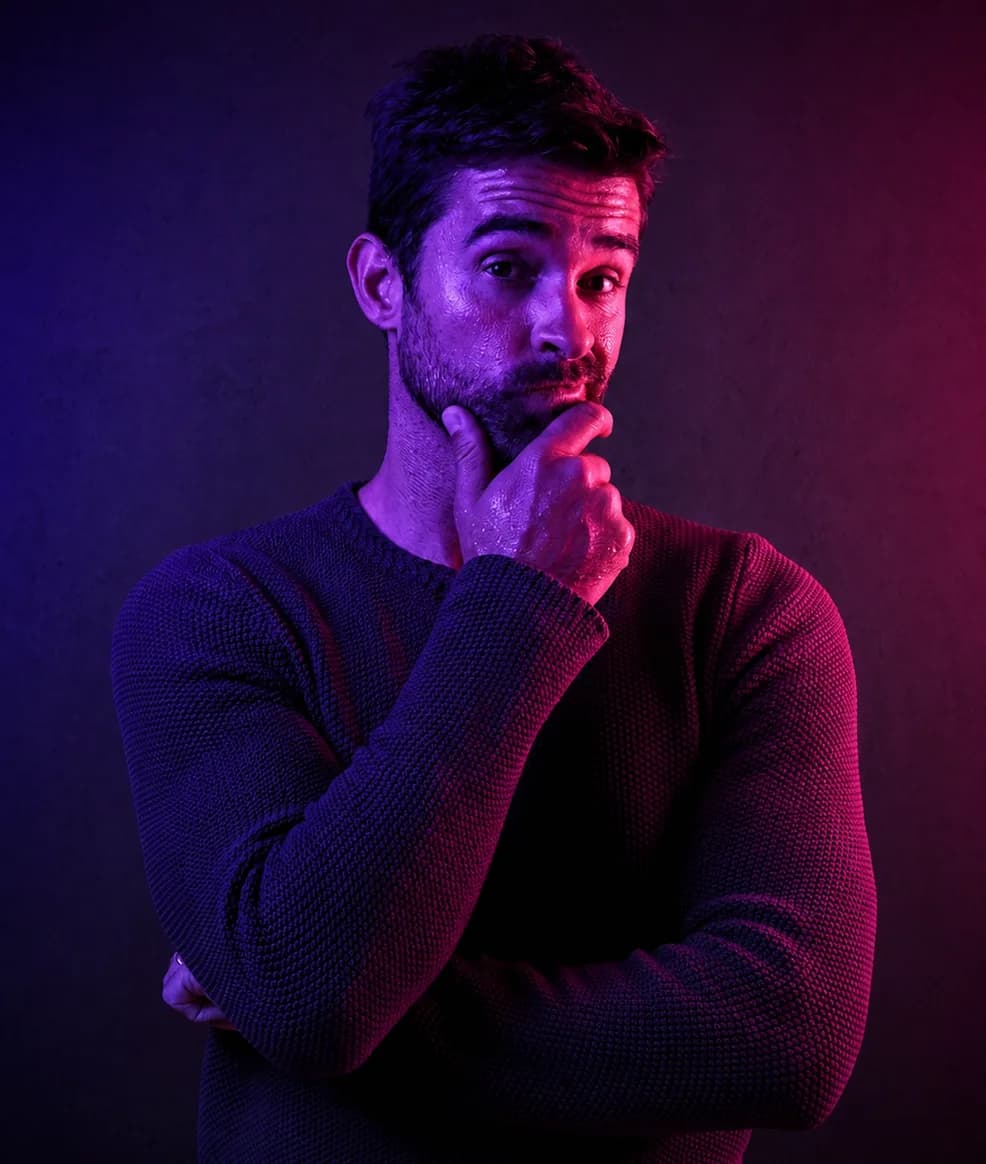 A man in a dark knit sweater, hand resting on his chin in thought, lit in purple and red.