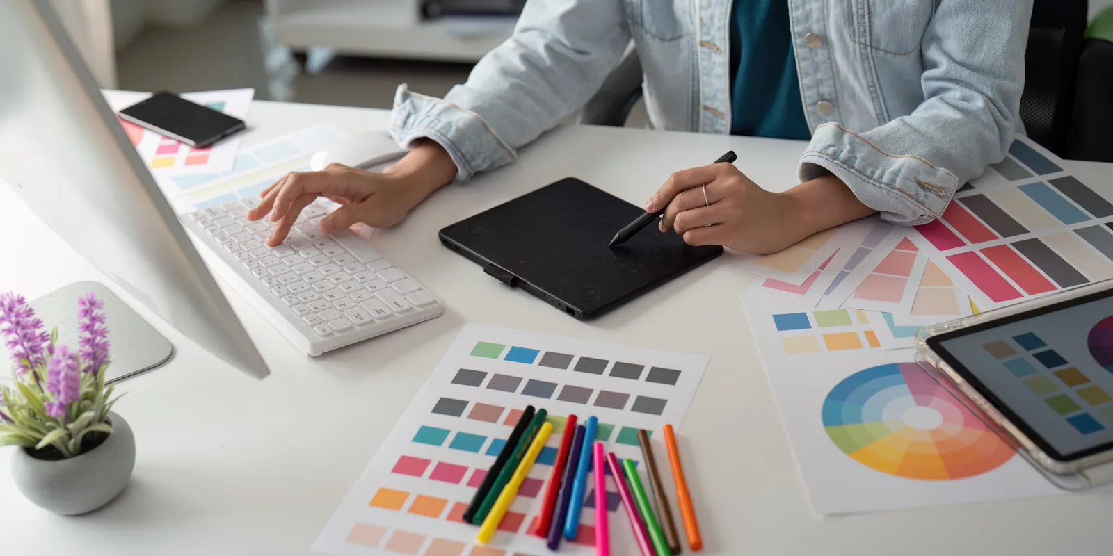 A designer working with a drawing tablet and stylus, surrounded by colour swatch sheets and marker pens.