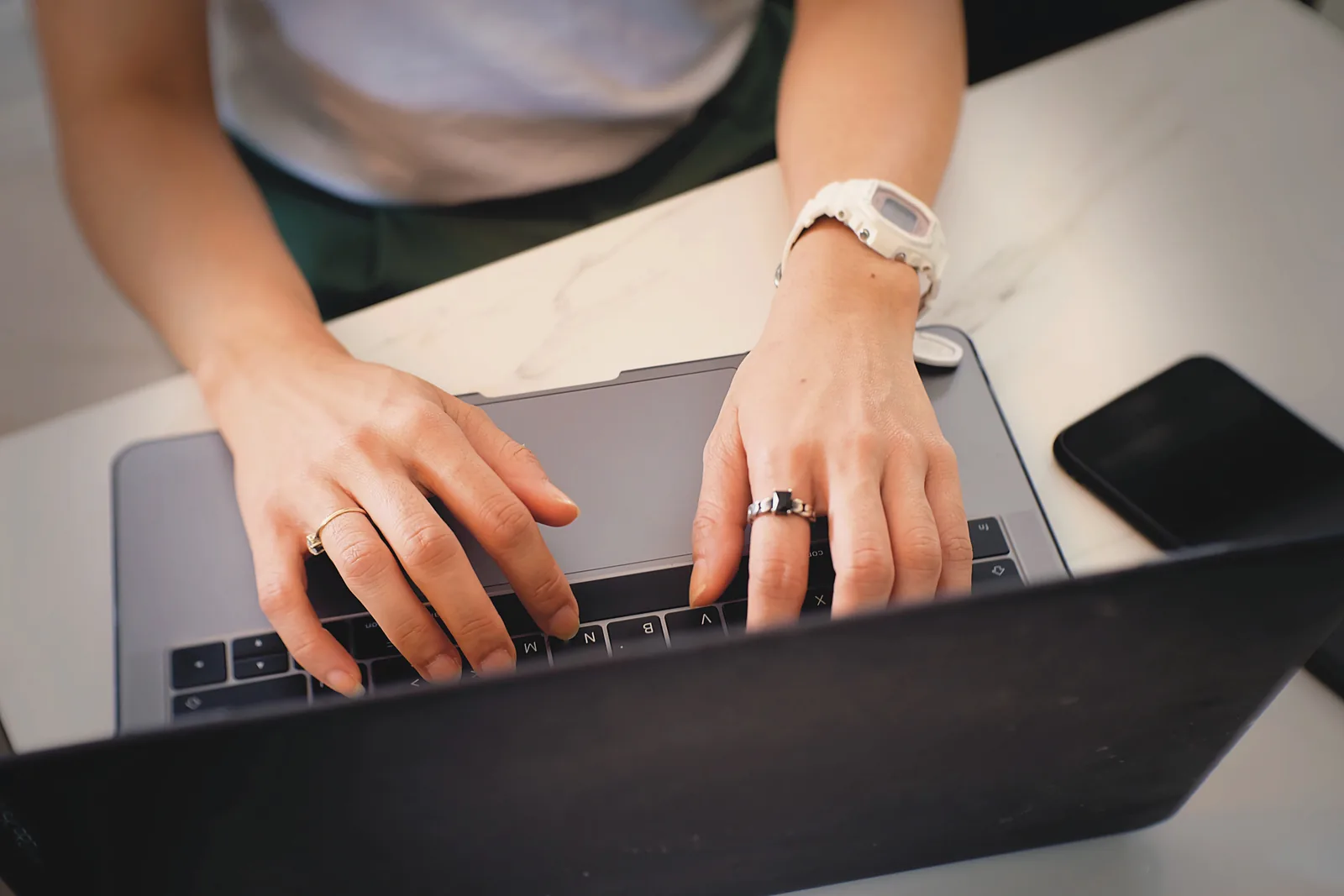 A woman typing on a laptop at a white marble table, a phone beside it.