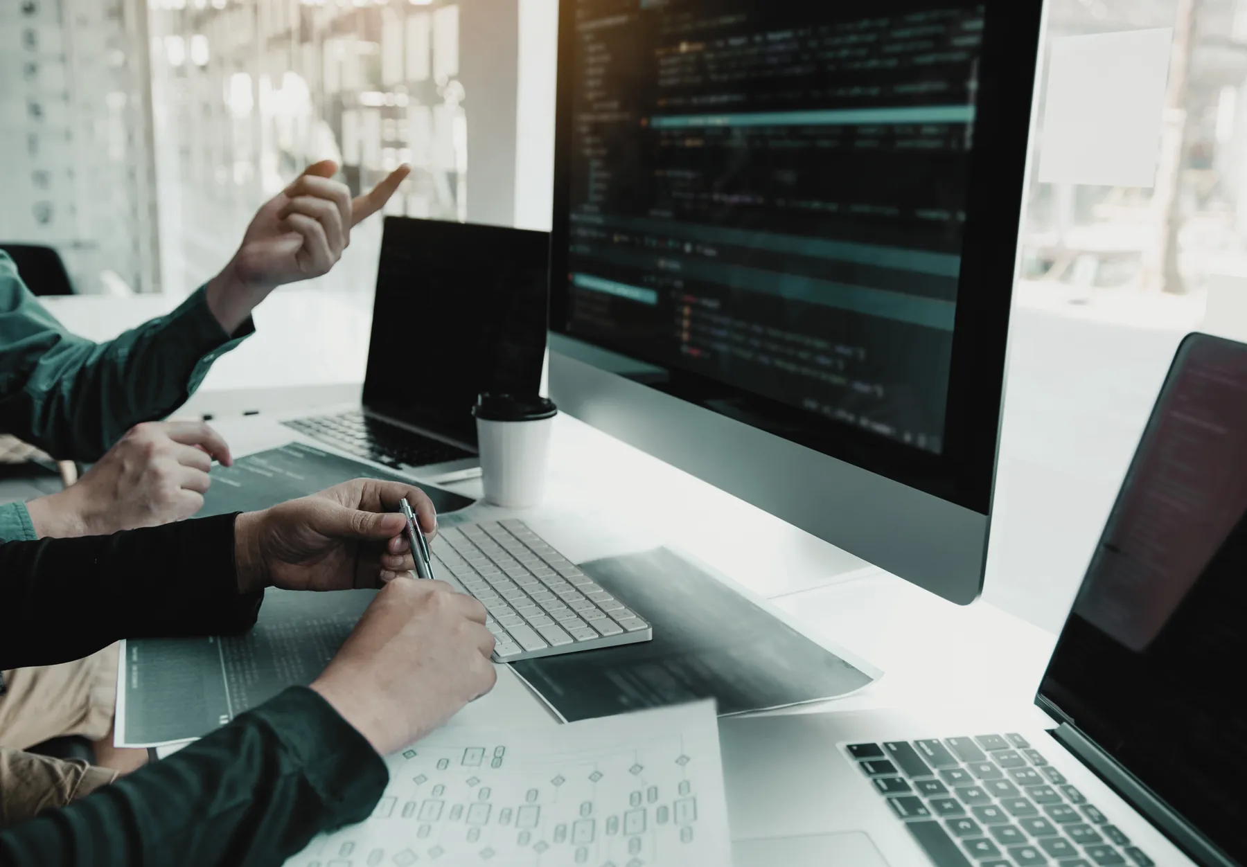 Developers pointing at code on a large monitor, with laptops and printed wireframes on the desk.
