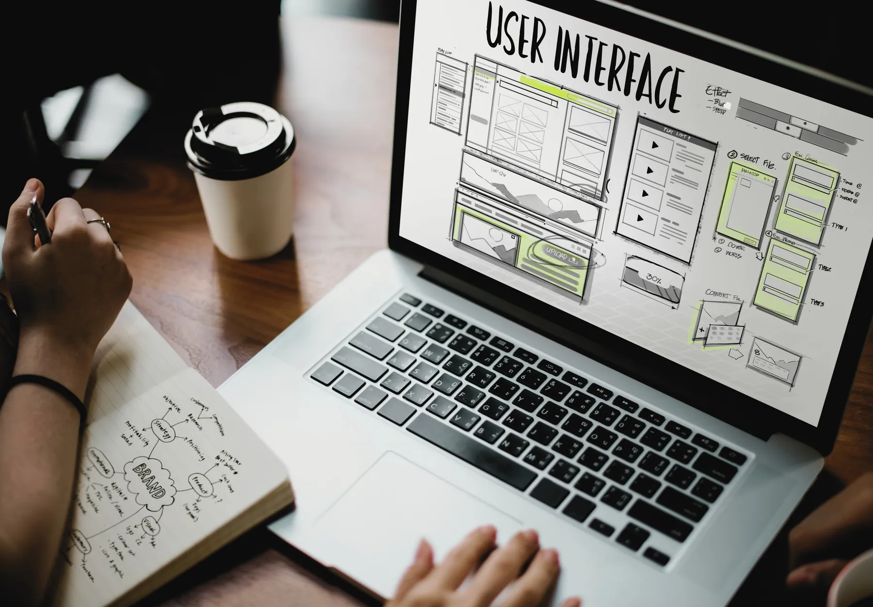 A laptop showing user interface wireframes, beside a notebook of sketched ideas and a coffee.