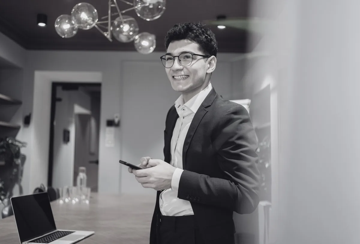 A smiling young entrepreneur in a blazer holding his phone in a modern office, a laptop open on the table beside him.