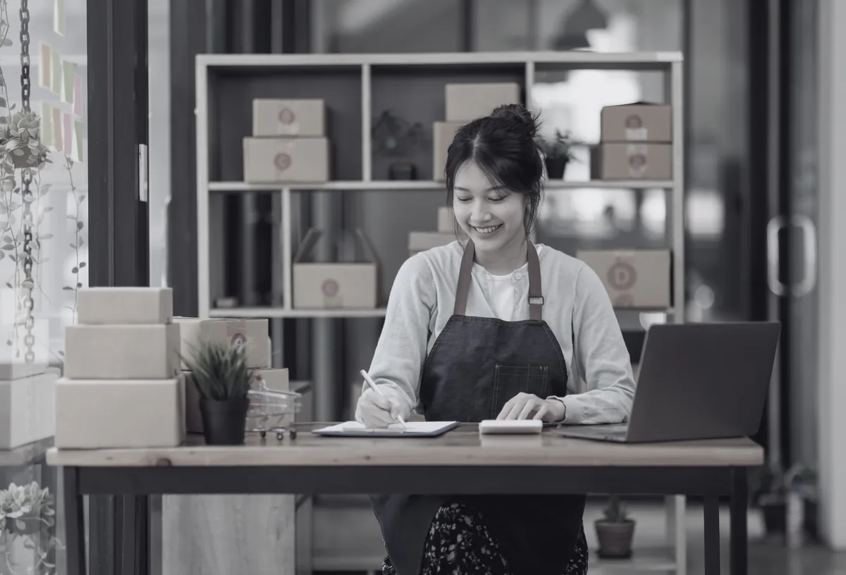 A smiling small business owner in an apron taking notes at her desk, shipping boxes and a laptop around her.